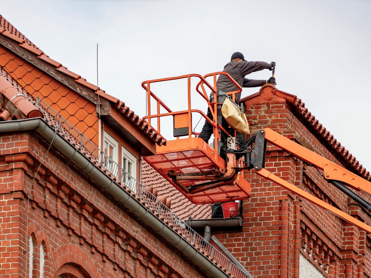 Roofer in a cherry picker conducting maintenance on a red brick building with a tiled roof.